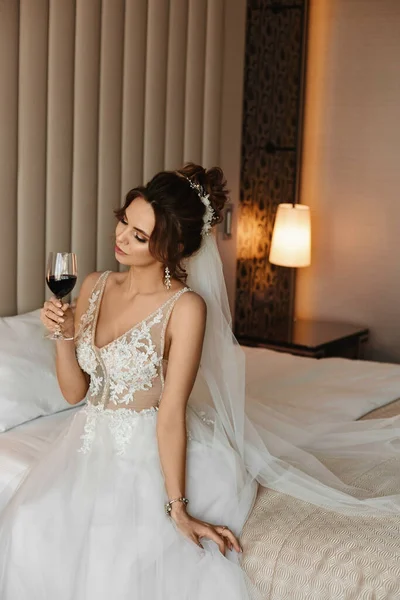 Mujer joven en un vestido de novia beber vino en la cama en un interior de lujo. Modelo chica con un peinado de boda en un vestido modish posando con una copa de vino. Concepto de moda de boda — Foto de Stock