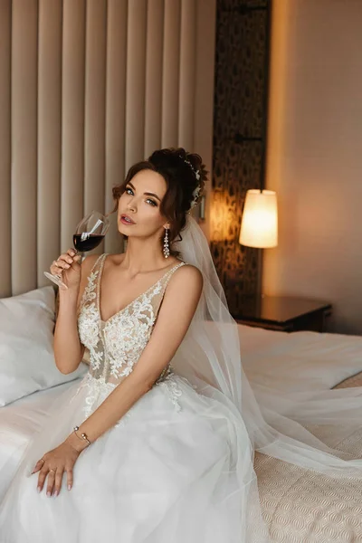 Jovem em um vestido de noiva bebendo vinho na cama em um interior luxuoso. Menina modelo com um penteado de casamento em um vestido modish posando com um copo de vinho. Conceito de moda de casamento — Fotografia de Stock
