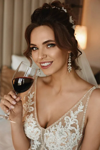 Ritratto di una bella ragazza modello in un abito da sposa bere vino sul letto in un interno lussuoso. Giovane sposa in abito modish posa con un bicchiere di vino. Concetto di moda nuziale . — Foto Stock