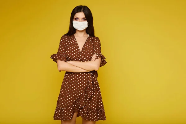 Mujer joven en vestido y máscara médica mirando a la cámara y posando sobre fondo amarillo, aislada. Chica modelo que se protege contra la epidemia de gripe, la alergia al polvo y el virus . — Foto de Stock