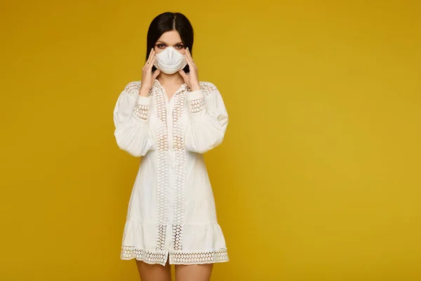 Model girl in a dress and a facial protective mask against virus and air pollution, isolated at the yellow background with copy space. Young woman wearing an anti-pollution face mask.