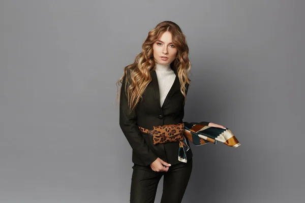 Молода жінка в модний костюм з пакетом з леопардовим поясом позує на сірому фоні, ізольована. Красива дівчина-модель у формальному одязі та з модним ременем з візерунком леопарда. Офіційна мода — стокове фото