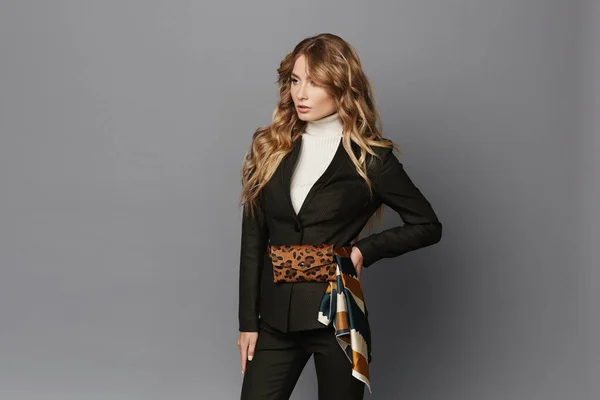 Young woman in modish suit with leopard belt bag posing at a grey background, isolated. Beautiful model girl in formal clothes and with trendy belt bag with leopard pattern. Spring fashion
