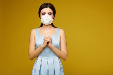 Beautiful young woman, in face mask and dress, with jointed arms and closed eyes prays at yellow background, isolated, copy space. Brunette girl in medicine mask prays for healing from epidemic of flu clipart