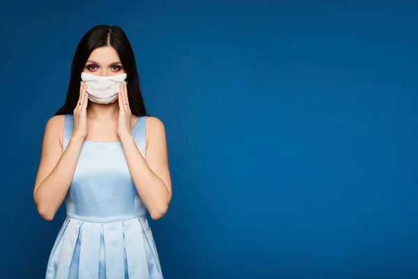 Mujer joven con vestido y máscara médica, mirando a la cámara, aislada sobre un fondo azul. Morena modelo chica en vestido y mascarilla protectora contra la epidemia de gripe, alergia al polvo . — Foto de Stock