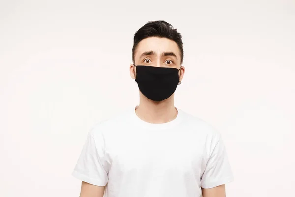 Наляканий чоловік у медичній масці дивиться в камеру. Наляканий молодий чоловік, боячись коронавірусу, носить білу футболку та захисну медичну маску проти вірусів, ізольований на білому тлі з місцем для тексту — стокове фото