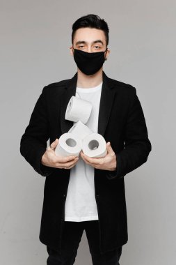 Siyah koruyucu maskeli yakışıklı bir genç. Elinde dört rulo tuvalet kağıdı var. Gri arka planda izole edilmiş. Koruyucu maskeli genç adamlar tuvalet kağıdı ile poz veriyorlar.