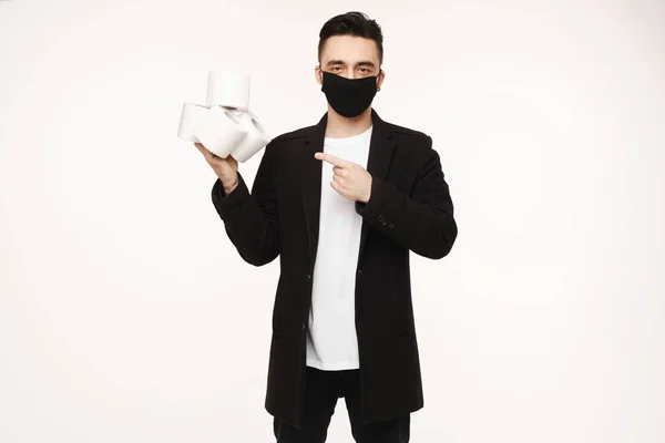 Красивий молодий чоловік в чорній захисній масці тримає чотири рулони туалетного паперу і вказує пальцем на нього, ізольовані на білому тлі. Молоді чоловіки в захисній масці для обличчя представляють туалетний папір — стокове фото