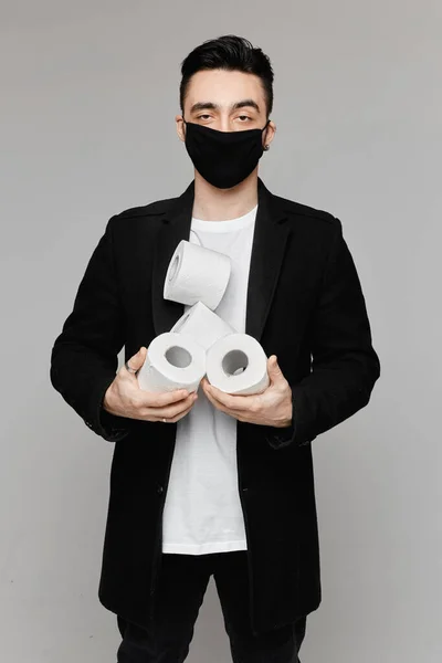 Красивий молодий чоловік в чорній захисній масці тримає чотири рулони туалетного паперу, ізольовані на сірому фоні. Молоді чоловіки в захисній масці для обличчя позує туалетним папером — стокове фото