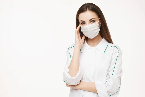 Krásná žena lékař nebo zdravotní sestra nosí ochrannou masku a lékařské šaty pózovat na bílém pozadí s kopírovacím prostorem, izolované. Mladá žena v lékařské uniformě. Koncept zdravotní péče — Stock fotografie