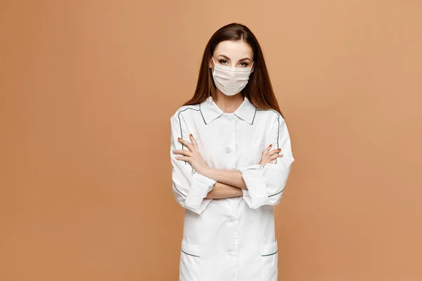 Médico femenino con máscara protectora posando sobre un fondo beige con espacio para copiar. Mujer joven en uniforme médico y máscara médica protectora aislada en el fondo beige, aislada — Foto de Stock