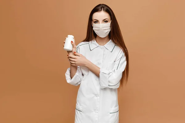 Young woman in medical clothes and protective mask posing with jar of pills, isolated at beige background. Female doctor keeps in hand pills. Virus and flu protection pills concept.