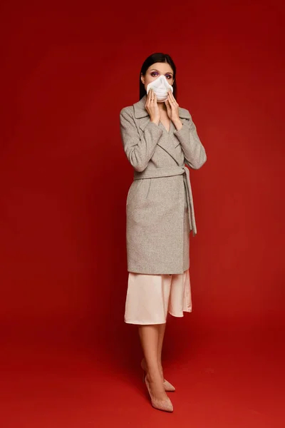 Hermosa mujer joven en abrigo y respirador posando en toda su longitud sobre fondo rojo con espacio para copiar, aislado. Modelo chica en máscara protectora y traje de primavera. Contaminación atmosférica y concepto de gripe estacional — Foto de Stock