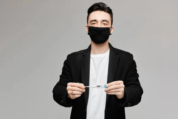 Молодий чоловік у чорній захисній масці тримає медичний термометр, ізольований на сірому фоні з копіювальним простором. Концепція здорового способу життя, сезонні хвороби та концепція сезонного грипу . — стокове фото