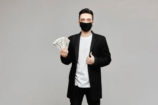 Красивий молодий чоловік в чорній захисній масці тримає пакет грошей, ізольований на сірому тлі з копіювальним простором. Концепція медико фармацевтичного бізнесу в сезоні грипу та пандемії — стокове фото