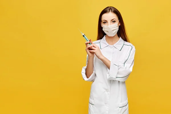 Mujer joven con ropa médica y máscara protectora posando con una jeringa, aislada en el fondo amarillo. Hermosa doctora lleva en la mano jeringa médica. Concepto de salud y farmacología — Foto de Stock