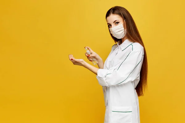 Una joven doctora con una bata blanca y una máscara protectora se pone un desinfectante en las manos. Mujer joven en uniforme médico pone antiséptico en las manos. Concepto de higiene . — Foto de Stock