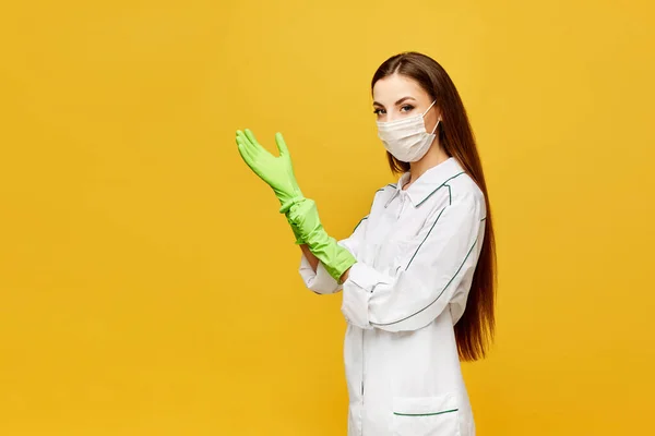 Doctora joven en uniforme blanco con máscara protectora y guantes aislados sobre fondo amarillo, espacio para copiar. Hermosa enfermera en bata blanca y máscara médica poniendo guantes médicos en sus manos — Foto de Stock