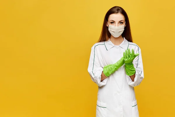 Doctora joven en uniforme blanco, máscara protectora y guantes aislados sobre fondo amarillo, espacio para copiar. Hermosa enfermera joven en bata blanca, máscara médica y guantes posando sobre fondo amarillo — Foto de Stock