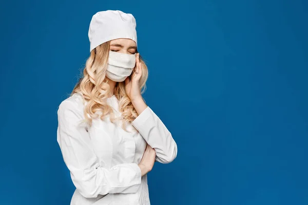 Doctora joven en bata blanca y máscara médica posando por un lado, aislada en un fondo azul con espacio para copiar. Enfermera en uniforme médico blanco y máscara protectora posando sobre fondo azul — Foto de Stock