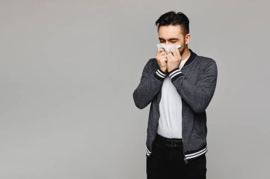 Hapşıran ve ağzı kağıt peçeteyle kaplanmış gri arka planda poz veren genç bir adam, izole edilmiş. Mendilli genç bir adamın stüdyo fotoğrafı. Soğuk mevsim konseptinde kötü adamlar. Kopyalama alanı oluştur