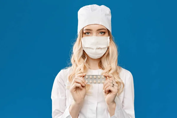 Mujer joven con abrigo médico blanco y máscara médica sostiene paquete de pastillas, aislado en el fondo azul con espacio para copias. Concepto de salud y prevención de enfermedades. Gripe estacional y virus — Foto de Stock