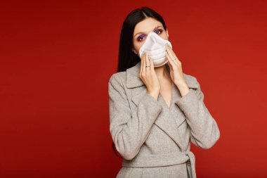 Ceketli ve solunum cihazlı genç bir kadın kameraya bakıyor ve kırmızı arka planda poz veriyor, izole edilmiş. Grip salgını, toz alerjisi ve hava kirliliğine karşı koruma kavramı
