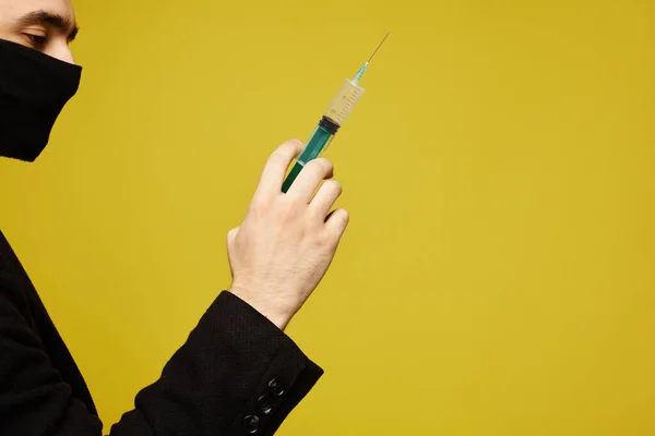 Side view of handsome man in black protective mask holding a syringe in his hand, isolated at the yellow background with copy space for your text.