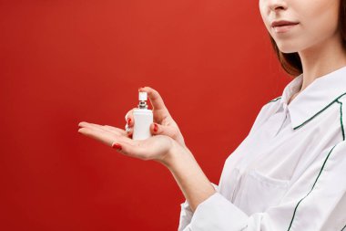 Tıp üniformalı genç bir kadın kırmızı arka planda dezenfektanla sprey kavanozu gösteriyor. Kopya alanı ile izole edilmiş. Kadın doktor avucunun içine ilaç sıkar.