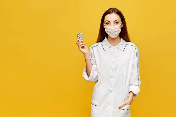 Doctora en uniforme y máscara médica protectora sosteniendo píldoras en la mano y posando en el fondo amarillo, espacio de copia, aislado. Concepto sanitario — Foto de Stock