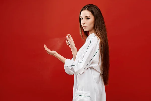 Mujer joven en uniforme médico rocía un desinfectante en la mano, aislado sobre un fondo rojo. La doctora usa aerosol desinfectante para desinfectar sus manos. Concepto de salud y medicina — Foto de Stock