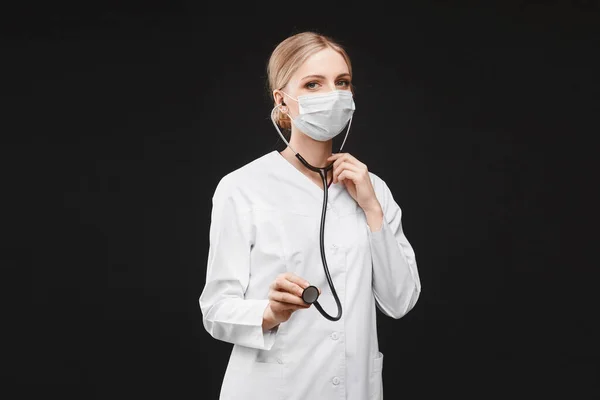 Mujer joven en uniforme médico y máscara protectora usando un estetoscopio, aislada en el fondo negro con espacio para copiar. Asistencia sanitaria y emergencia — Foto de Stock