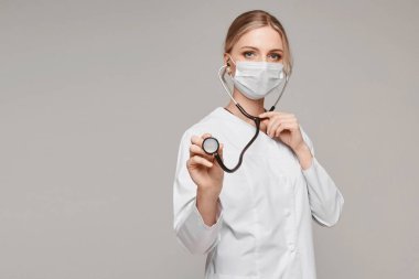 Tıbbi üniformalı ve koruyucu maskeli genç bir kadın steteskop kullanıyor ve gri arka planda poz veriyor, izole. Sağlık sistemi ve acil durum kavramı