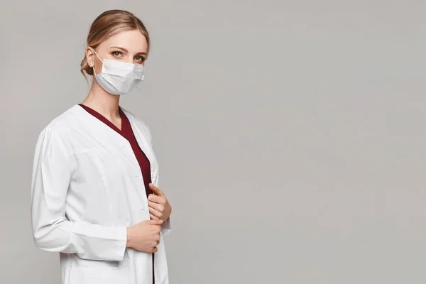 Beautiful female doctor or nurse wearing protective mask and medical gown posing on grey background with copy space, isolated. Young woman in medical uniform. Healthcare system concept — Stock Photo, Image