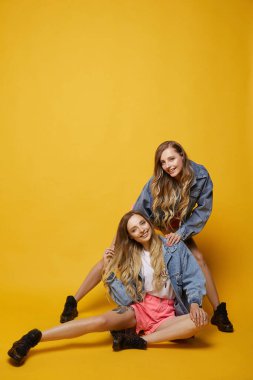 Kot ceketli, pembe şortlu, ince vücutlu iki güzel genç ikiz kadın sarı arka planda poz veriyorlar.