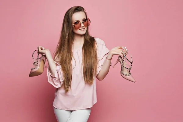 Mulher loira jovem com maquiagem brilhante, em blusa bege e óculos de sol rosa na moda com sapatos elegantes em suas mãos, posando em fundo rosa em estúdio — Fotografia de Stock