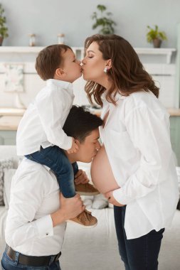 Küçük çocuk, mutlu babası hamile karısının karnını öperken annesini öpüyor. Mutlu aile konforlu bir evde vakit geçiriyor. Aynı kıyafetlerin içinde neşeli bir aile.