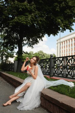 Gelinlikli mutlu genç gelin şehir parkında çıplak ayakla oturur ve patates kızartması yer. Düğün modası kavramı