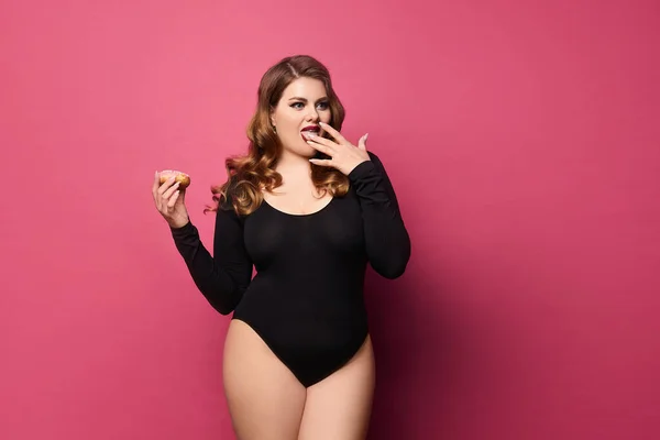 Plus Size Model Girl im schwarzen Body hält süße Donuts in der Hand und leckt ihre Finger, isoliert auf rosa Hintergrund mit Kopierraum. Konzept der xxxl Mode — Stockfoto