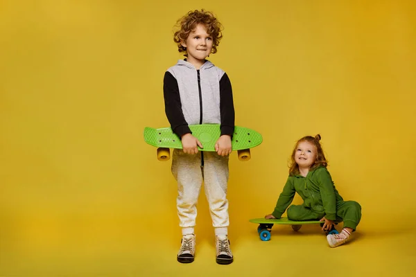 Niños con estilo en trajes modernos posando con patinetas. Niña y niño aislados en el fondo amarillo con espacio de copia. Moda infantil — Foto de Stock