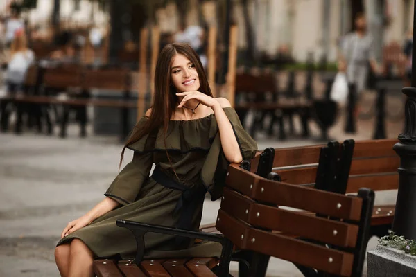 Fille modèle dans une robe est assis sur le banc brun dans la ville par une journée d'été ensoleillée. Style de vie en plein air portrait de mode . — Photo