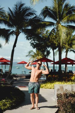 Yaz günü tropik otellerde genç bir atletik adam kaslı gövdesini kameraya bakarak gösteriyor. Yaz tatili ve seyahat