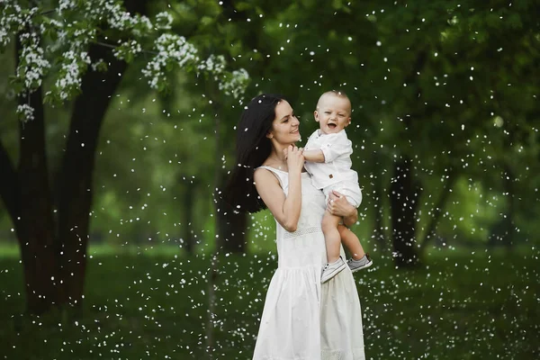 Hermosa mujer joven en un vestido blanco largo con un lindo niño pequeño en camisa y pantalones cortos en sus manos posando en el jardín verde en el día de verano —  Fotos de Stock