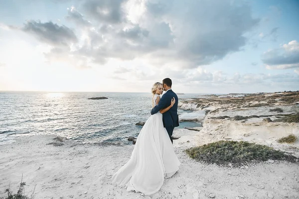 Молодая пара обнимается на белых скалах на побережье Кипра. Невеста в свадебном платье жених в костюме позирует с красивым пейзажем на заднем плане — стоковое фото
