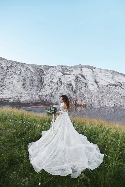 Incredibile giovane sposa in abito da sposa di lusso con un bouquet da sposa su un prato vicino a montagne bianche. Concetto di moda matrimonio. Cerimonia nuziale all'aperto . — Foto Stock
