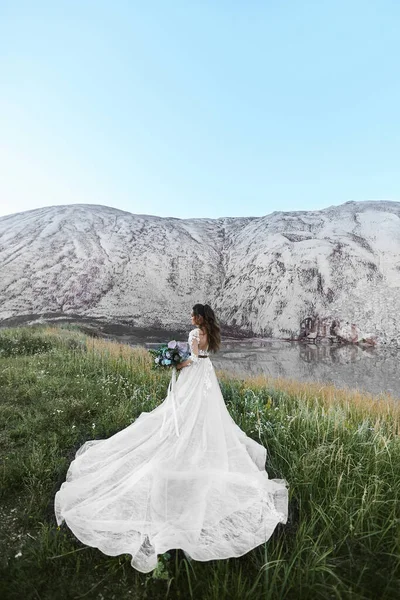 Giovane donna in abito da sposa con un bouquet da sposa in posa con la schiena in un bellissimo paesaggio. Concetto di moda matrimonio. Cerimonia nuziale all'aperto . — Foto Stock