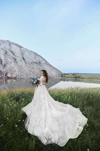 Schöne junge Frau in einem Brautkleid mit Brautstrauß und posiert vor einer wunderschönen Landschaft. Hochzeitsmodekonzept. — Stockfoto