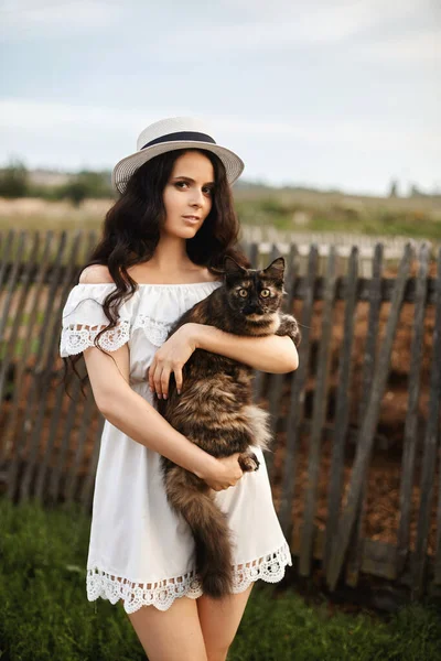 Young woman in short white dress posing with big cat on her hands outdoors. Beautiful model girl in short white dress holding a cat on her hands and posing in a rural background. Summertime — Stock Photo, Image