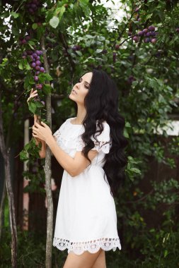 Beyaz yaz elbisesi içindeki yumuşak kız erik ağacının yanında poz veriyor ve yan bakıyor. Yaz bahçesinde kısa beyaz elbiseli esmer bir kadın. Yaz zamanı.