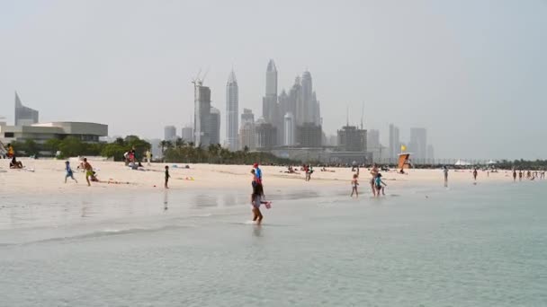 Gyerekek úszkálnak a tengerben egy homokos strandon Szufouh hátterében felhőkarcolók Dubai, Egyesült Arab Emírségek. Tiszta napsütéses nap 2020. március 16. — Stock videók